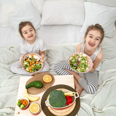 Healthy food at home. Happy two cute children eating fruits and vegetables in the bedroom on the bed. Healthy food for children and teenagers.